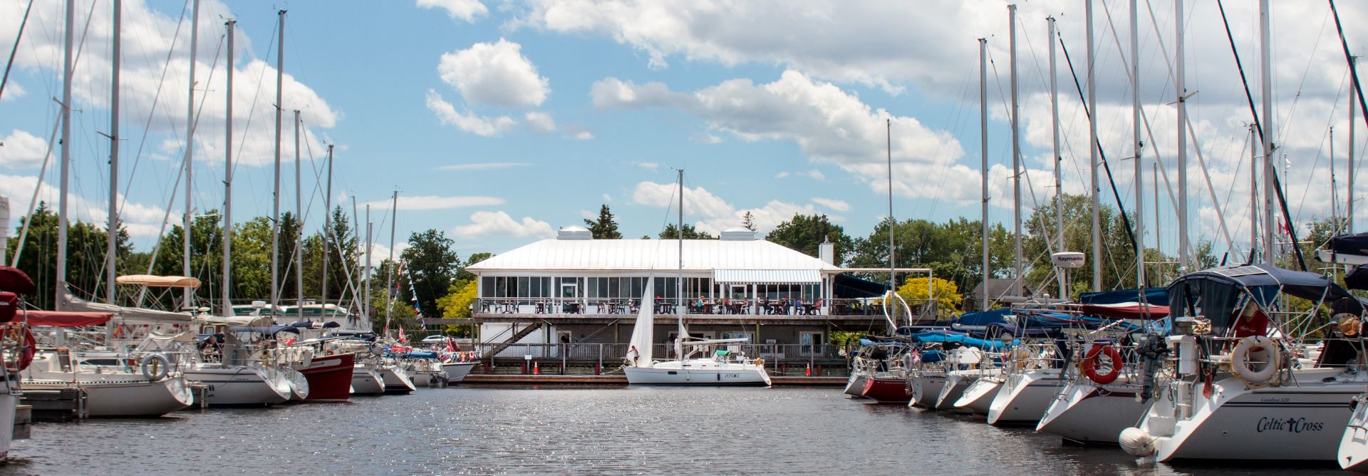 A view of the club from the water on a beautiful summer day with rows of boats framing the photo and a foresail partially hoisted on a large boat at the service dock